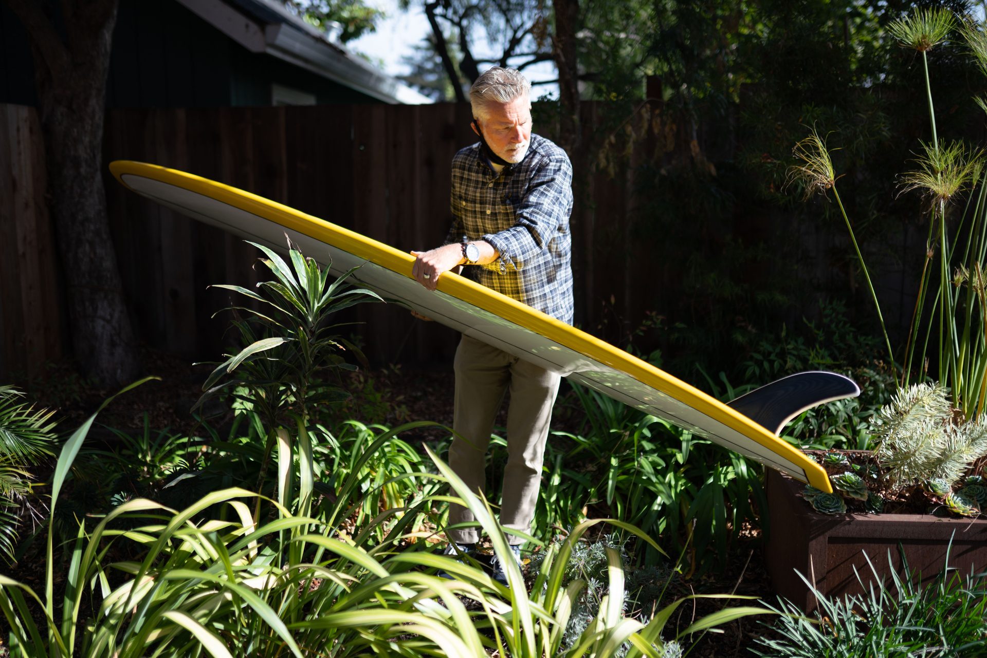 A man carrying a yellow longboard through a garden